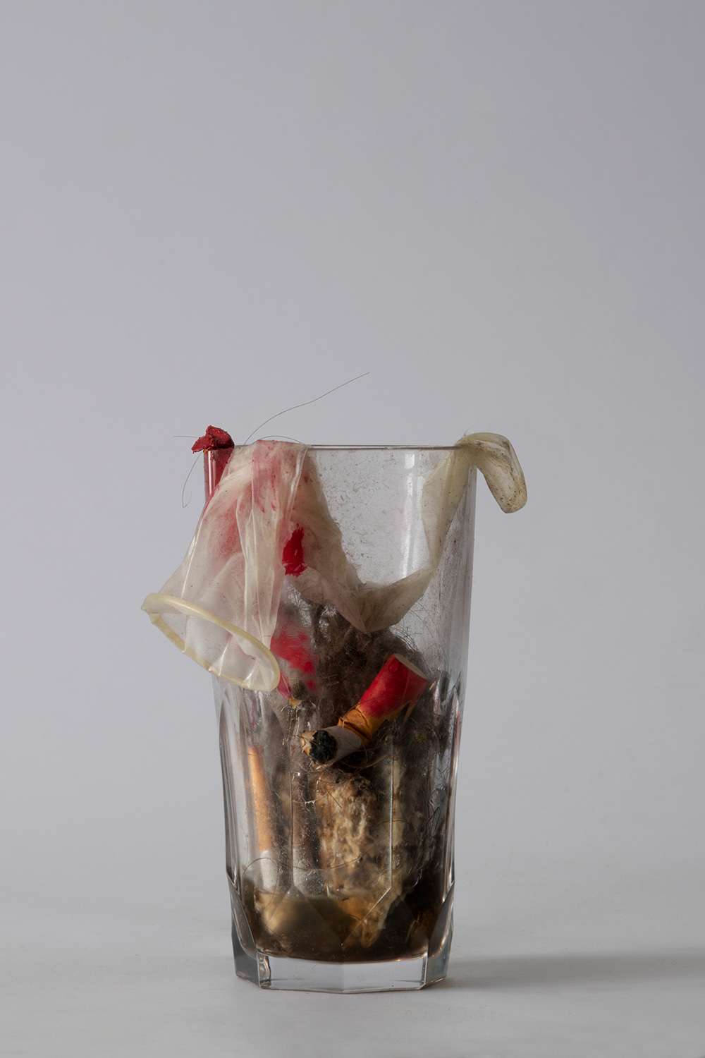 Glass full of cigs butts, condom, lipstick and dust on grey background
